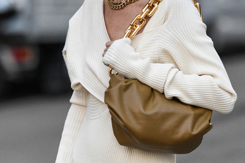 kobieta w swetrowej białej sukience niesie brązową torbę na ramieniu, torba jest na złotym łańcuchu