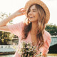 uśmiechnięta kobieta w różowej sukience, słomkowym kapeluszu i trzymająca kwiaty